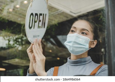 Asian Small Business Owner Woman Wearing Protection Face Mask Turning Open Sign Board On Glass Door For Reopening Cafe Restaurant After Coronavirus (covid-19) Quarantine. Food And Drink Concept