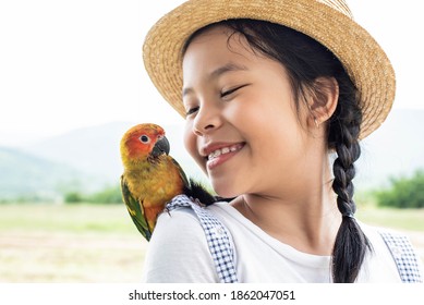 Asian small cute child is playing a colourful small bird, bird is standing on her shoulder. Cute smiling girl playing with her pet green Sun Conure parrot. Exotic pet.