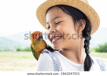 Asian small cute child is playing a colourful small bird, bird is standing on her shoulder. Cute smiling girl playing with her pet green Sun Conure parrot. Exotic pet.