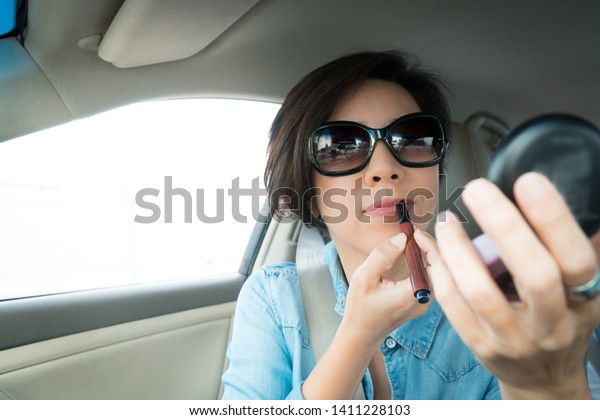 An asian smart\
looking woman applying her make-up in the car while driving on the\
way to work in rush hour. Middle aged woman, Put on lipstick,\
Driving in the city concept.