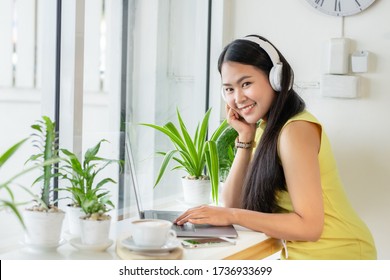 Asian Smiling Girl Student Wear Wireless Headphone Study Online With Skype Teacher In A Coffee Shop, Woman Learn Language Listen Lecture Look At The Camera Sit In Cafe, Distant Education, New Normal