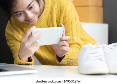 Asian Smiling Girl Using Smartphone To Shoot White Sneaker Posted For Sale On The Internet.startup Small Business Owner Working With Tablet At Workplace.happy Owner & Seller Check Product To Customer.