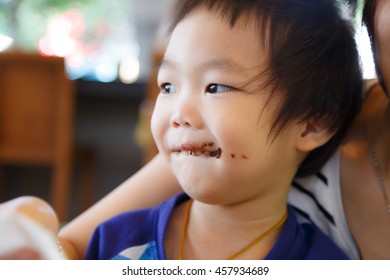 Asian Smiling Kid Eating Chocolate At Coffee Shop With His Mom. Smeared Stained With Chocolate Lips