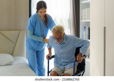 Asian smiling nurse helping senior man get out of bed nursing care support patient while getting out of bed and moving to wheelchair at home helping handicapped elderly stand up