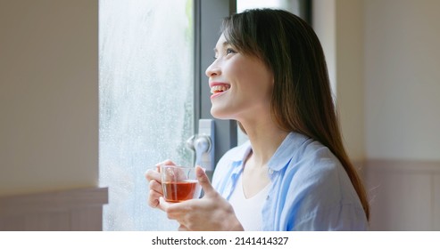 Asian Smiling Young Woman Is Drinking Tea While Looking Through Window On A Rainy Day At Home