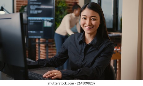 Asian Software Developer Smiling And Resuming Work Typing Code On Computer Keyboard Sitting At Desk In Big Data Office. Database Admin Working Casually In Artificial Intelligence Development Agency.