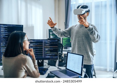 Asian software developer wearing a virtual reality headset works on a VR project, with a colleague focused on her computer