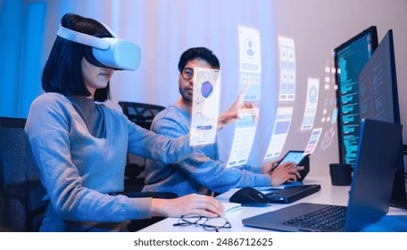 Asian software developer wearing a virtual reality headset works on a VR project, with a colleague focused on her computer