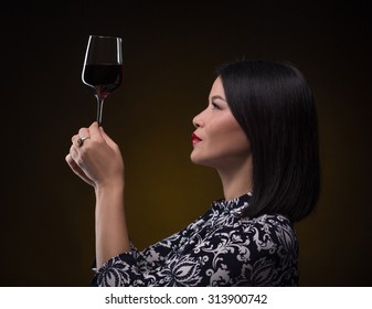 Asian Sommelier Woman Looking At Glass Of Red Wine Very Attentively. Beautiful Lady With Blach Hair Holding Glass In Front Of Her.