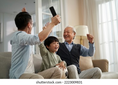 asian son father grandfather sitting on couch at home watching live broadcasting of football match on TV together - Powered by Shutterstock