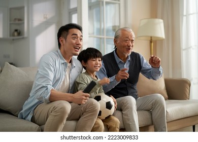 asian son father grandfather sitting on couch at home watching live broadcasting of football match on TV together - Powered by Shutterstock