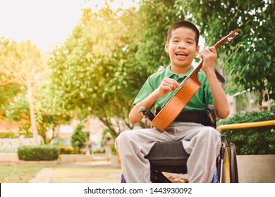 Asian Special Child On Wheelchair Is Playing Ukulele Happily On The Playground, Nature Background, Life In The Education Age Of Disabled Children, Happy Disabled Kid Concept.