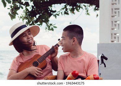 Asian Special Child On Wheelchair Is Singing, Playing Music Therapy On The Beach With Parent,Natural Sea Beach Background,Life In The Education Age Of Disabled Children,Happy Disability Kid Concept.