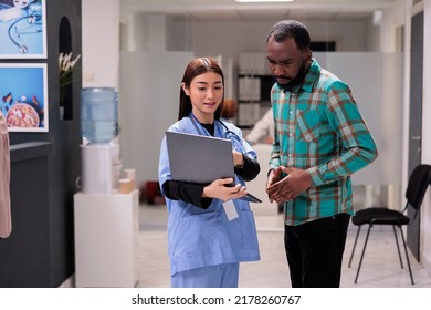 Asian Specialist Consulting African American Man Using Laptop To Show Disease Diagnosis, Explaining Healthcare Service In Hospital Reception Lobby. Diverse People Doing Checkup Exam At Facility.