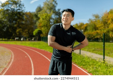 Asian sportsman has severe pain in back and abdominal muscles, man peed training in stadium on sunny day massaging side of back, athlete on jogging pulled muscle.