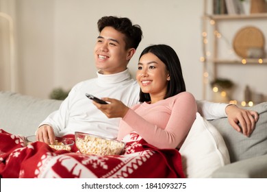 Asian Spouses Enjoying Christmas Eve Watching TV And Xmas Movies Sitting On Couch In Living Room At Home. Asian Family Watches Television Indoors. New Year And X-Mas Celebration Concept