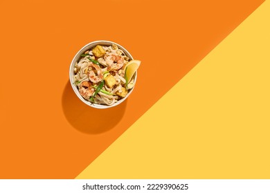Asian street food - pad thai noodles with shrimp on coloured background. Pad thai udon with prawns and lemon in bowl on orange and yellow backdrop. Trendy concept udon noodles with seafood