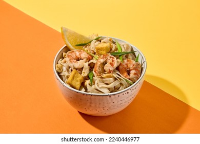 Asian street food - pad thai noodles with shrimp on coloured background. Pad thai udon with prawns and lemon in bowl on orange and yellow backdrop. Trendy concept udon noodles with seafood