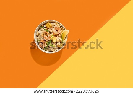 Asian street food - pad thai noodles with shrimp on coloured background. Pad thai udon with prawns and lemon in bowl on orange and yellow backdrop. Trendy concept udon noodles with seafood
