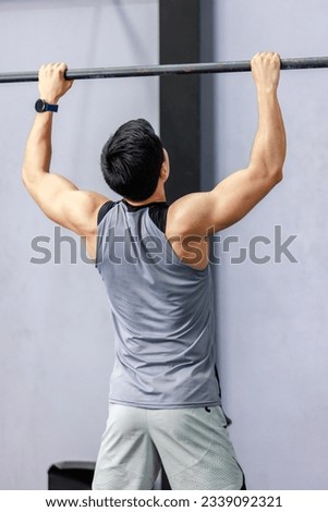 Asian strong unrecognizable unknown young shirtless male muscular fitness model in sporty shorts holding pulling up gymnastic rings lifting working out back muscle strength in gym on gray background.