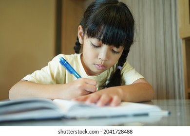 Asian Student Child Girl Writing On The Paper Book. Asian Genius Student Doing Homework At Home. Education, Study, And Learning Concept.         