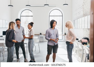 Asian Student In Shorts Listening Charming Blonde University Mate In Campus. Pretty African Female Office Worker Telling Colleagues Something Funny And Laughing.