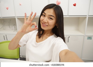 Asian Student Showing Okay Sign To Camera While Making Selfies In Library. Happy Brunette Lady Posing On Selfie In Library Or Office.