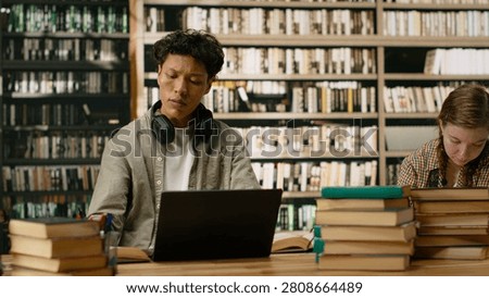 Asian student using headphones and taking notes with laptop and open book, studying at library table near stack of books, study session with textbooks, academic workspace.