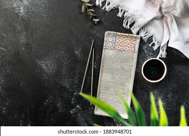 Asian Style Table Setting With Long Ceramic Plate, Sushi Chopsticks And Bowl With Soy Sauce On Black Rustic Background With Bright Palm Leaves And White Napkin, Copy Space For Your Design.