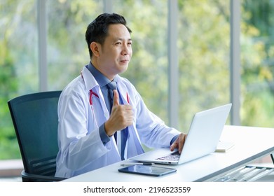 Asian Successful Professional Middle Aged Male Doctor In White Lab Coat With Stethoscope Sitting Using Laptop Notebook Computer Working Researching Online In Glass Windows Hospital Clinical Office.