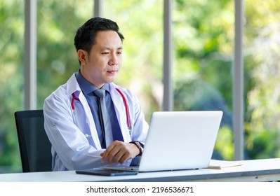 Asian Successful Professional Middle Aged Male Doctor In White Lab Coat With Stethoscope Sitting Using Laptop Notebook Computer Working Researching Online In Glass Windows Hospital Clinical Office.