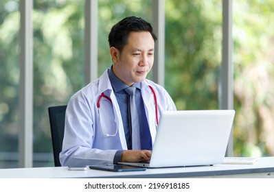 Asian Successful Professional Middle Aged Male Doctor In White Lab Coat With Stethoscope Sitting Using Laptop Notebook Computer Working Researching Online In Glass Windows Hospital Clinical Office.