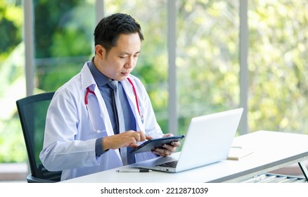 Asian Successful Professional Middle Aged Male Doctor In White Lab Coat With Stethoscope Sitting Using Laptop Notebook Computer Working Researching Online In Glass Windows Hospital Clinical Office.