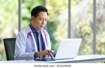 Asian Successful Professional Middle Aged Male Doctor In White Lab Coat With Stethoscope Sitting Using Laptop Notebook Computer Working Researching Online In Glass Windows Hospital Clinical Office.