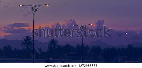 Asian sunset at road\
with lamps street in silhouette tone. Urban street, car and\
lampposts on the\
background.
