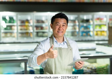 Asian Supermarket Worker Looks Into The Camera And Smiles Shows Thumbs Up, Inspects The Quality Of The Goods Uses A Tablet Near The Freezers