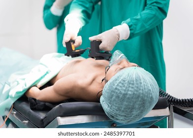 Asian surgical team in green scrubs using defibrillator on patient in operating room. Focus on emergency heart failure, professional teamwork quick precision response, healthcare concept in hospital - Powered by Shutterstock