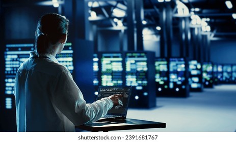Asian system administrator running code and programming on laptop, troubleshooting and upgrading server farm equipment designed to accommodate mainframes, networking racks and storage arrays