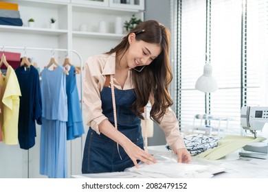 Asian Tailor Woman Talking On Phone With Customer In Tailoring Atelier. Attractive Beautiful Young Female Fashion Designer Working And Checking New Fashionable Handmade Clothes Collection In Workshop.