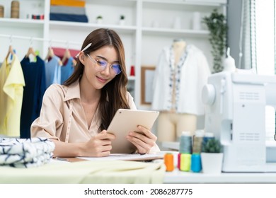 Asian tailor woman working to design new clothes in tailoring atelier. Attractive young female fashion designer dressmaker use tablet to find reference to think and design new clothes in workshop room