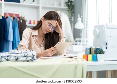 Asian Tailor Woman Working To Design New Clothes In Tailoring Atelier. Attractive Young Female Fashion Designer Dressmaker Use Tablet To Find Reference To Think And Design New Clothes In Workshop Room