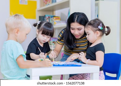 Asian Teacher Helping Her Students To Painting On Paper While Studying In The Kindergarten