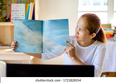 Asian Teacher Online Learning.Female Showing Story Book. Kindergarten And Preschool Reading Lesson Through Laptop Because Of Corona Virus Lockdown. Homeschooling Through Video Call.