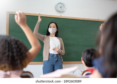 Asian Teacher Wearing Protective Face Mask And Children Or Schoolkids Raising Hands In Classroom While Covid-19 Pandemic. New Normal Life Concept.