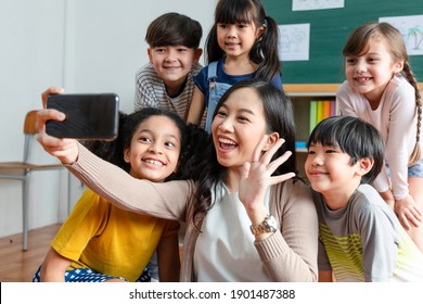 Asian Teachers Use Smartphones To Take Selfies With A Group Of Deversity Elementary School Students In The Classroom. They Use A Facetime Phone. Talk To Friends