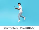 Asian Teen Boy Holding Laptop Jumping And Browsing Internet Posing Over Blue Studio Background. Side View Shot Of Student Guy Using Computer In Mid Air. Technology And Gadgets