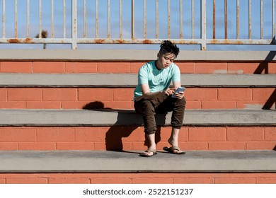 Asian teen boy sitting outdoor wearing blue shirt listening to music and using a smartphone under the sun. Sunny day. Asian teen boy chilling outdoor. Asian teen boy looks calm and thoughtful. - Powered by Shutterstock