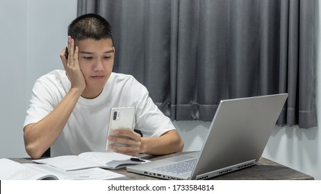 Asian Teen Boy Is Using Mobile Stay Online Connecting To The Internet While Doing Homework With A Notebook, Book, And Laptop On Desk At Night In The Bedroom. Youth Man As Students Uses A Phone.