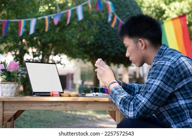 Asian teen gay holds pink color bottle and read details on table, learning to draw in the park which decorated with rainbow flag and LGBTQ+ flagline, concept for special LGBT people events.