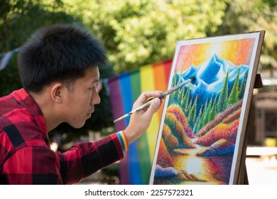 Asian teen gay in plaid shirt wears rainbow wristband, holding paintbrush to check his painting in park which decorated with rainbow flags, concept for LGBT people activity around the world.